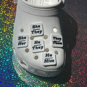 May include: White clogs with multiple square-shaped charms attached. The charms have black text that reads "She/They", "She/Her", "He/They", "They/Them", and "He/Him".