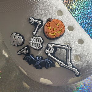 May include: White shoe with Halloween-themed charms. The charms include a pumpkin, a bat, a skull, and a skeleton.