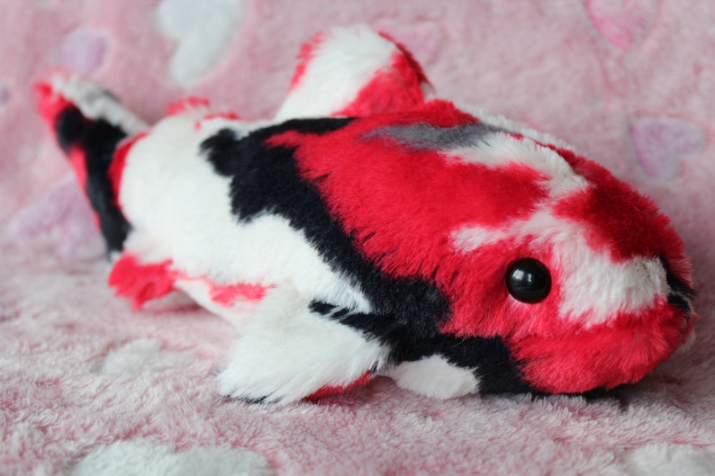 Puede incluir: Un juguete de peluche con forma de pez koi en rojo, negro y blanco. El animal de peluche tiene un cuerpo redondeado, aletas y un peque&ntilde;o ojo negro. El juguete est&aacute; hecho de material suave y esponjoso y est&aacute; sobre una superficie rosa.