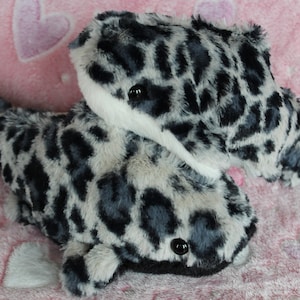 May include: Two plush toys shaped like whales with a black and white leopard print. The whales are lying on a pink blanket with white hearts.