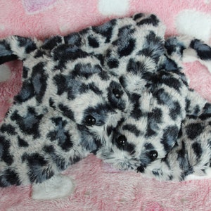 May include: A plush toy shaped like a snow leopard with a black and white spotted pattern. The toy is lying on a pink fuzzy surface.