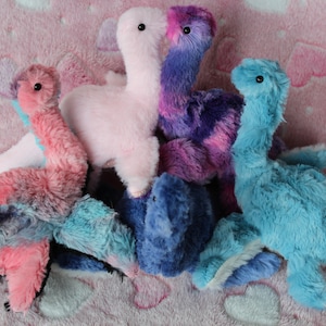 Op de afbeelding: Vijf pluche dinosaurus speelgoed in verschillende kleuren, waaronder roze, paars, blauw en teal. De dinosaurussen zijn op een roze en witte pluche deken gestapeld.
