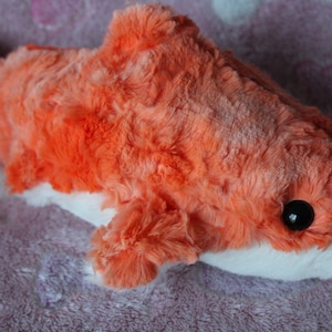 May include: A plush orange and white shark toy with a black eye. The shark is made of soft, fuzzy fabric and has a white belly.