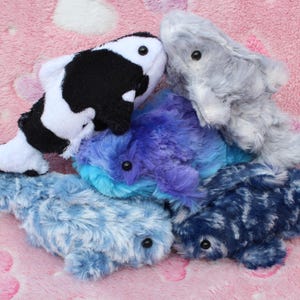 Può includere: Una collezione di pesci di peluche in vari colori, tra cui bianco e nero, grigio, blu e viola. I giocattoli sono disposti su una superficie rosa con motivo a cuore, creando una scena giocosa e stravagante.