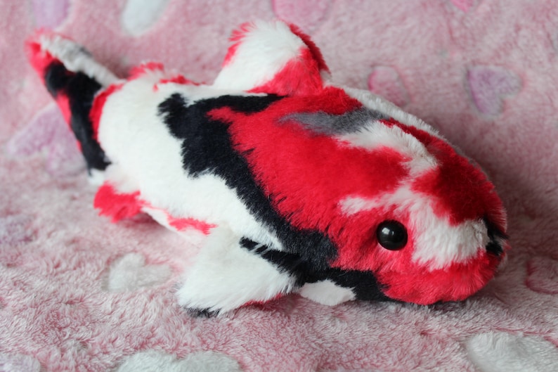 Puede incluir: Un juguete de peluche con forma de pez koi con un vibrante patr&oacute;n de rojo, negro, blanco y gris. El juguete blando presenta un cuerpo redondeado, aletas y un peque&ntilde;o ojo negro. Se encuentra sobre un fondo rosa con estampado de corazones.
