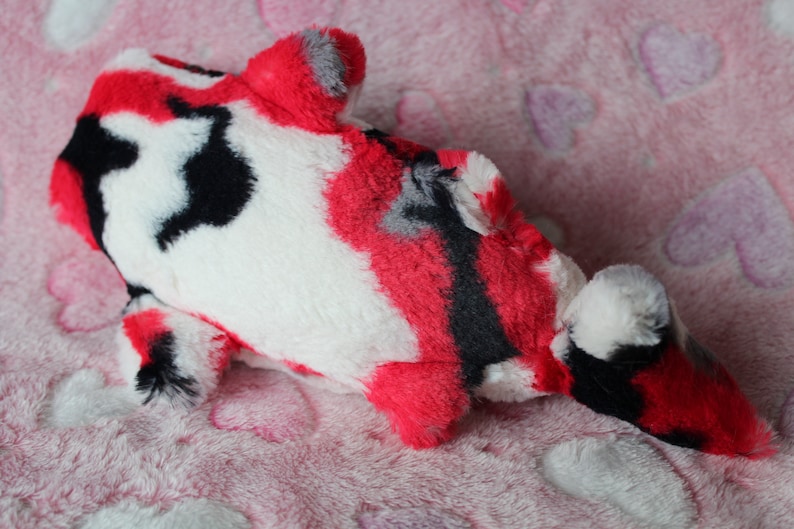 Puede incluir: Un juguete de peluche con forma de pez koi en rojo, negro, blanco y gris. El juguete blando tiene un dise&ntilde;o estampado y est&aacute; sobre una superficie rosa con estampado de corazones. El juguete est&aacute; dise&ntilde;ado para parecerse a un pez koi.