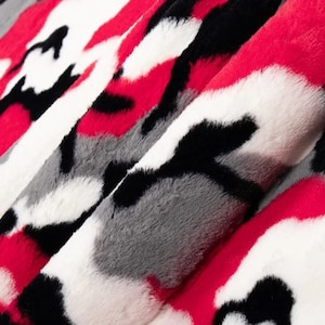 Puede incluir: Primer plano de una tela estampada con un dise&ntilde;o de camuflaje. El textil presenta una mezcla de colores rojo, negro, blanco y gris. El material suave y afelpado est&aacute; doblado, revelando la textura y las variaciones de color.