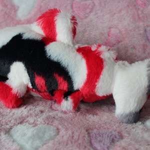 Puede incluir: Un juguete de peluche con forma de pez koi en rojo, blanco, negro y gris. El peluche tiene un aspecto suave y texturizado y est&aacute; sobre una superficie rosa con patrones de coraz&oacute;n. El juguete est&aacute; dise&ntilde;ado para parecerse a un pez koi.