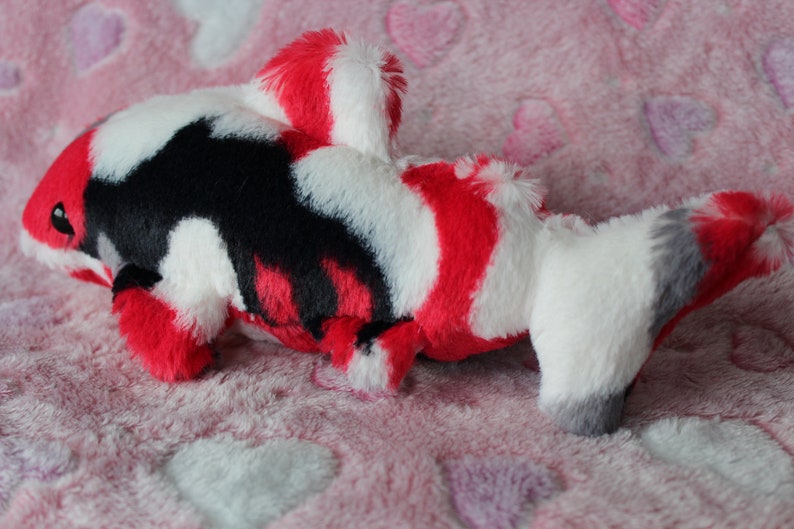 Puede incluir: Un juguete de peluche con forma de pez koi en rojo, blanco, negro y gris. El peluche tiene un aspecto suave y texturizado y est&aacute; sobre una superficie rosa con patrones de coraz&oacute;n. El juguete est&aacute; dise&ntilde;ado para parecerse a un pez koi.