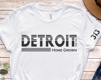 May include: White t-shirt with black text that reads "DETROIT EST 1701 HOME GROWN" with the number "313" on the sleeve. The t-shirt is laid out on a wood background with a pair of brown sandals, blue jeans, and a brown fedora hat.