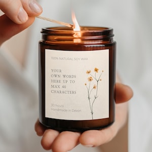 May include: A lit candle in an amber glass jar with a cream-coloured label featuring a botanical illustration of small yellow flowers. The label text reads: 100% NATURAL SOY WAX, YOUR OWN WORDS HERE UP TO MAX 40 CHARACTERS, 30 hours, Handmade in Devon.