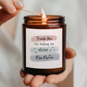 May include: A lit candle in an amber glass jar with a white label featuring three horizontal watercolour brush strokes in dusty pink, sage green, and slate blue. The text reads Thank You for helping me shine Miss Martin with star accents.