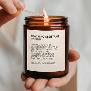 May include: A brown glass jar candle with a cream-coloured label featuring black text that reads: TEACHING ASSISTANT [TA'] NOUN. INSPIRES YOU TO BE BETTER. GUIDES YOU WHEN YOU ARE LOST. A SKILLED ENCOURAGER AND WONDERFUL MENTOR. CHANGES THE WORLD, ONE CHILD AT A TIME. SEE ALSO; INSERTNAME.