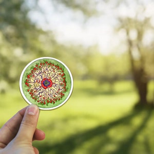 May include: A round sticker with a colorful, intricate design of a flower with red, yellow, and green petals. The center of the flower is purple and white.