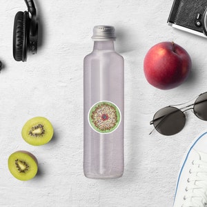 May include: A clear glass water bottle with a silver lid and a round sticker with a colorful mandala design. The bottle is surrounded by various items, including headphones, sunglasses, a kiwi fruit, an apple, and a pair of white sneakers.