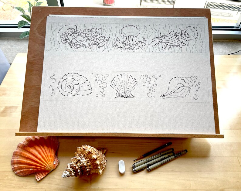 以下が含まれることがあります： イーゼルの上に置かれた白い紙に、貝殻とクラゲの絵が描かれている。絵は黒いインクで描かれており、巻貝、ホタテガイ、巻貝、クラゲ3匹が描かれている。貝殻とクラゲの周りに、泡を表す小さな円も描かれている。