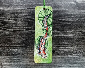 May include: A hand-painted wooden bookmark featuring a colorful koi fish swimming through green lily pads. The bookmark is a vibrant and detailed illustration.