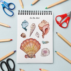 Puede incluir: Una libreta blanca con encuadernación en espiral y las palabras "To Do List" escritas en la portada. La libreta está rodeada de lápices de colores y tijeras. La libreta tiene ilustraciones de conchas marinas en varios colores y formas.