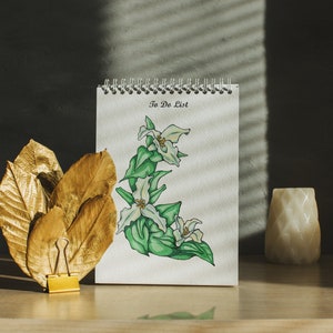 May include: A white notepad with a spiral binding and the words "To Do List" written on the front. The notepad is decorated with a hand-drawn illustration of white flowers with green leaves. The notepad is sitting on a wooden surface with gold-painted leaves and a small white candle holder.
