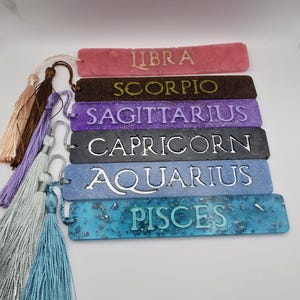 May include: Six rectangular resin bookmarks featuring the zodiac signs Libra, Scorpio, Sagittarius, Capricorn, Aquarius, and Pisces. Each bookmark has a unique color, embedded glitter or foil accents, raised lettering, and a matching fabric tassel.