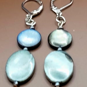 May include: A pair of dangle earrings with silver-tone lever-back closures. Each earring features two iridescent, oval-shaped beads in shades of blue and green. The earrings are displayed against a brown background.
