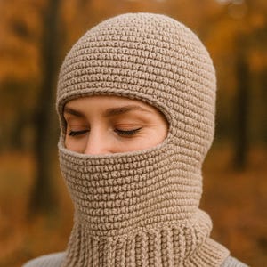 May include: A beige crochet balaclava with a ribbed neck and face opening. The head covering is made of a textured yarn and is worn by a person with their eyes closed. The background is blurred with autumn colors.