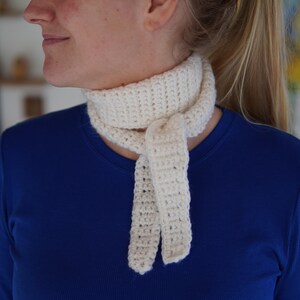 Puede incluir: Bufanda de crochet color crema atada al cuello. La bufanda está hecha de un hilo grueso con una puntada texturizada. La persona de la imagen lleva una camiseta azul.