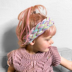 May include: A young person with pink hair wearing a multi-colored braided headband. The headband is made of yarn in shades of pink, green, and purple.