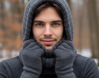 May include: A person wearing a gray knitted hood, scarf, and gloves set. The hood is pulled up, framing the face. The gloves are covering the hands. The background is blurred, with trees visible.