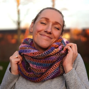 May include: A woman wearing a colorful crocheted infinity scarf. The scarf is made with a variety of shades of purple, orange, brown, and blue yarn.