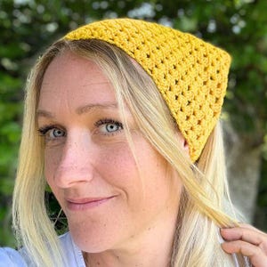 May include: A yellow crochet headband with a square shape. The headband is made of a textured, open-weave fabric. The person in the image has blonde hair and blue eyes.