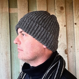 May include: A dark gray, ribbed knit beanie hat. The hat is made of a soft, textured yarn and has a folded brim. The hat is worn by a person. The person is wearing a black jacket and a black and white striped scarf.