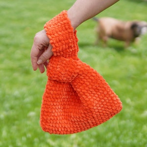 Può includere: Una borsa di tessuto arancione brillante e peloso con un manico annodato. La borsa è realizzata in un materiale morbido e testurizzato.