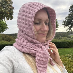 Puede incluir: Una capucha de ganchillo en color rosa empolvado con textura acanalada que cubre la cabeza y envuelve el cuello. La prenda presenta extremos largos y puntiagudos que se atan en la parte delantera, creando un aspecto superpuesto con un patrón uniforme.