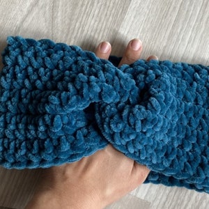 May include: A teal blue crocheted headband with a twisted knot design. The headband is made with a soft, fuzzy yarn.