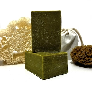 以下が含まれることがあります： オリーブグリーンの長方形の石鹸が2つ積み重ねられ、ヘチマ、小さな巾着袋、茶色のスポンジが背景にあります。石鹸はざらざらした表面をしています。アイテムは白い背景に配置されており、スパやバス製品をテーマにしています。