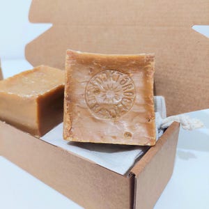 Pode incluir: Duas barras de sabão castanho, uma estampada com um desenho circular, repousam numa caixa de cartão castanha forrada com um tecido branco. O sabão tem um aspeto rústico e natural. A caixa está aberta, revelando o sabão.
