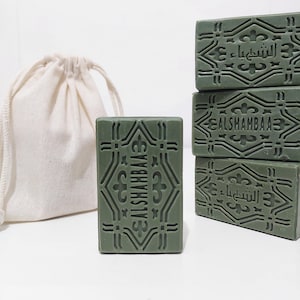 May include: A stack of four green soap bars with the text "ALSHAHBAA3" and "الشيحبا" printed on them. The soap bars are sitting on a white surface next to a white drawstring bag.