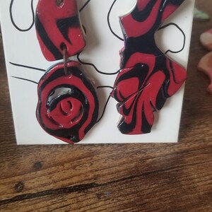 May include: A pair of dangle earrings with red and black marbled clay. The earrings are made of two pieces each, one larger and one smaller. The larger piece is shaped like a leaf and the smaller piece is round.