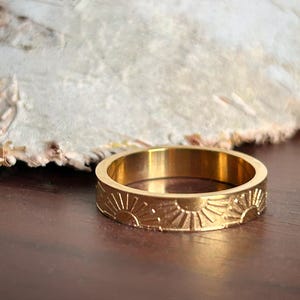 May include: A gold-toned ring with a repeating sun design. The ring is made of stainless steel and has a polished finish.