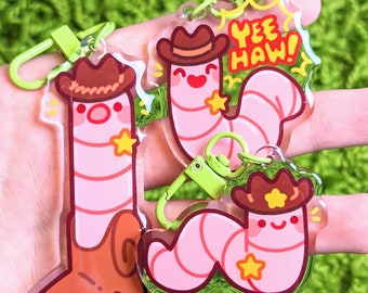 Puede incluir: Dos llaveros de gusano rosa con sombreros de vaquero y una estrella amarilla. Los llaveros están unidos a llaveros verdes con anillos de metal. Uno de los llaveros tiene el texto "Yee Haw!"
