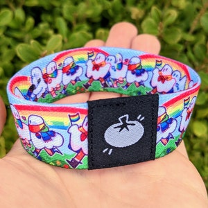 May include: A colorful woven wristband with a rainbow design and cartoon ghosts wearing scarves. The wristband has a black adjustable closure with a white tomato logo.