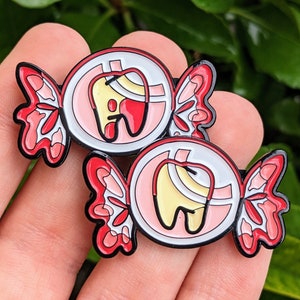 Könnte beinhalten: Zwei Emaille-Pins in Form von Süßigkeiten mit einem Zahnmotiv. Die Süßigkeit ist rosa und weiß mit einem roten und gelben Zahn darin. Die Pins sind an einer Hand befestigt.