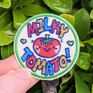 May include: A white, round embroidered patch with a green border. The patch features a cartoon tomato with a smiling face and the text "MILKY TOMATO!" in colourful, outlined letters.