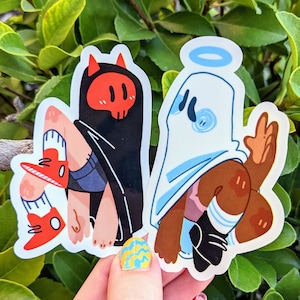 Könnte beinhalten: Zwei Vinyl-Aufkleber mit Cartoon-Figuren. Einer ist ein Teufel in einem schwarzen Umhang mit rotem Totenkopfgesicht, der andere ein Engelgeist mit blauem Heiligenschein. Beide Aufkleber haben einen weißen Hintergrund und werden vor grünem Laub gehalten.