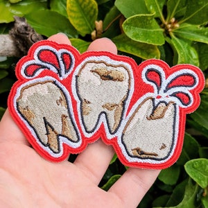 May include: Embroidered patch featuring three teeth with decay, set against a red background with stylized blood droplets. The teeth are off-white with brown and black detailing, outlined in black and white.