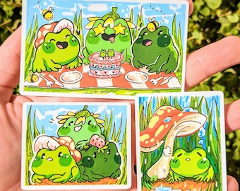 May include: Three sticker designs featuring cartoon frogs in various settings. The first sticker shows three frogs having a picnic with a cake and a bee. The second sticker shows three frogs sitting under a mushroom. The third sticker shows a frog sitting under a mushroom with raindrops falling.