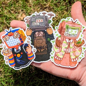 May include: Three colorful stickers featuring cartoon characters with television screens for heads. The characters are wearing different outfits and have different expressions. The stickers are on a hand.