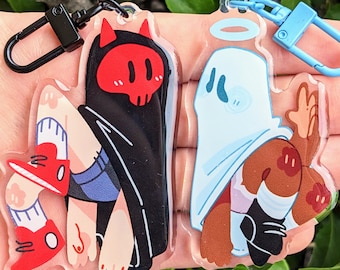 May include: Two acrylic keychains featuring cartoon characters. The first keychain features a red-headed character wearing a black cloak and red shoes. The second keychain features a blue-headed character wearing a white sheet and brown shoes.