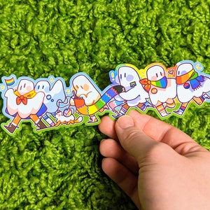 May include: A sticker featuring a line of seven cartoon ghosts wearing rainbow-colored clothing and accessories. The ghosts are holding hands and walking together. The sticker is on a green background.